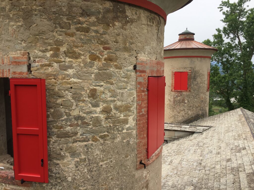 Scuri in alluminio rosso su misura installati sulle aperture di una torre in pietra, con tetti e paesaggio collinare sullo sfondo.
