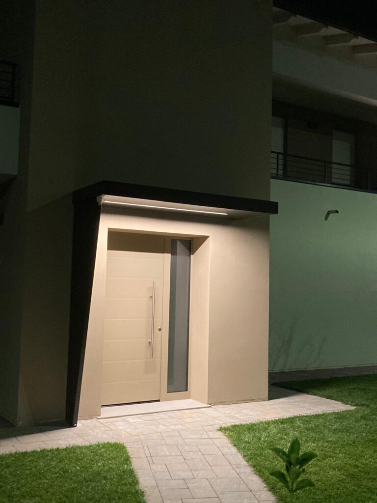 Porta d’ingresso moderna color avorio con lunga maniglia verticale e fasce laterali vetrate, inserita in una facciata contemporanea e illuminata nelle ore serali.