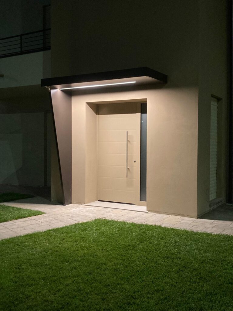 Porta d’ingresso moderna color avorio con lunga maniglia verticale e fasce laterali vetrate, inserita in una facciata contemporanea e illuminata nelle ore serali.